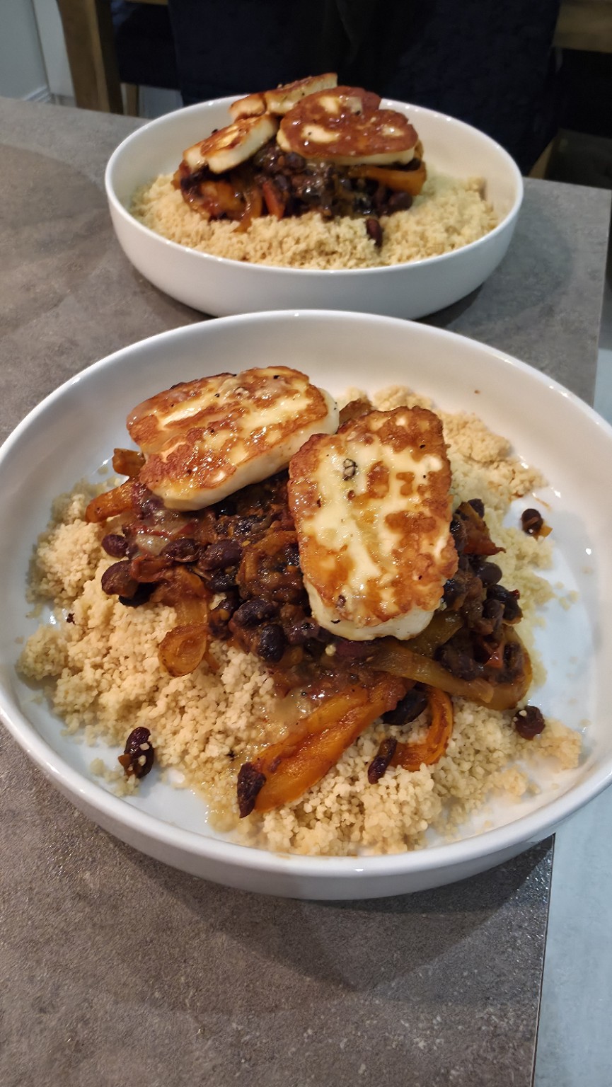Two bowls of couscous topped with smoky black bean peppers and golden halloumi, served with a glass of white wine