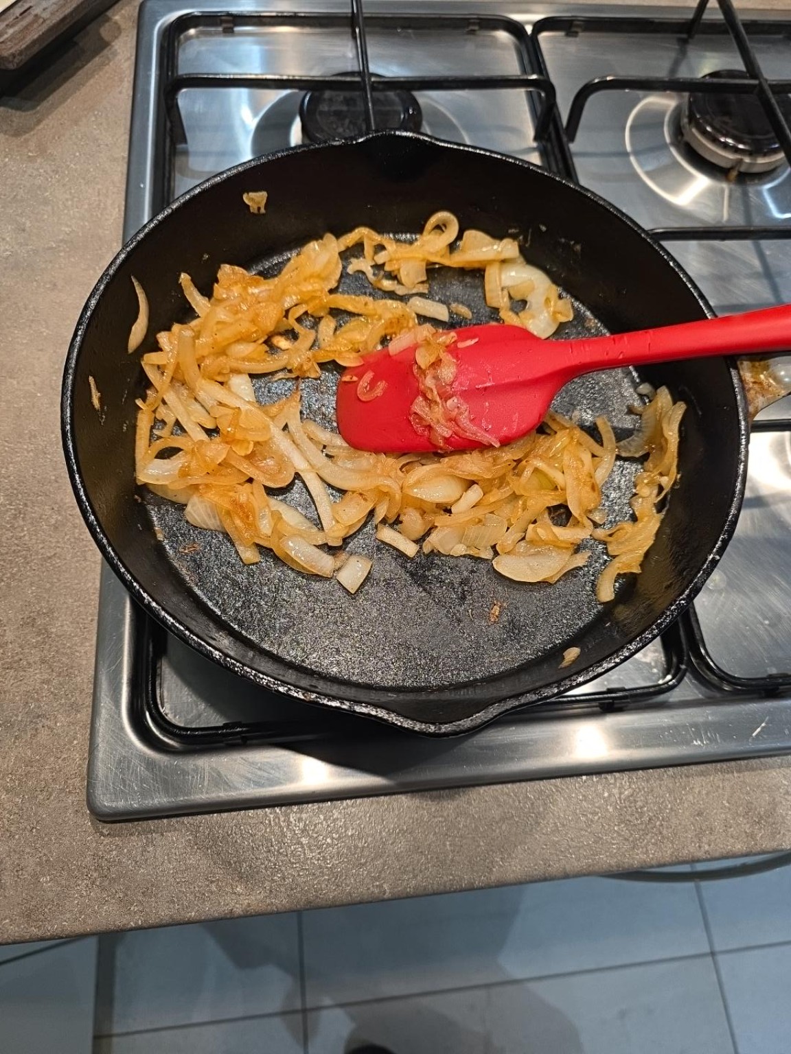 Thinly sliced onions turning golden in a cast-iron pan alongside a red spatula