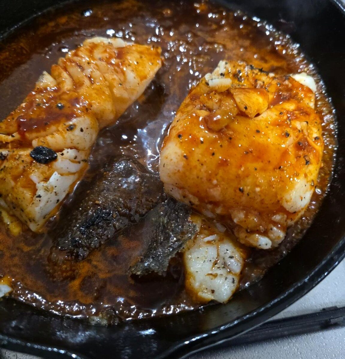 Two portions of cod coated in reddish glaze searing in a black cast-iron frying pan