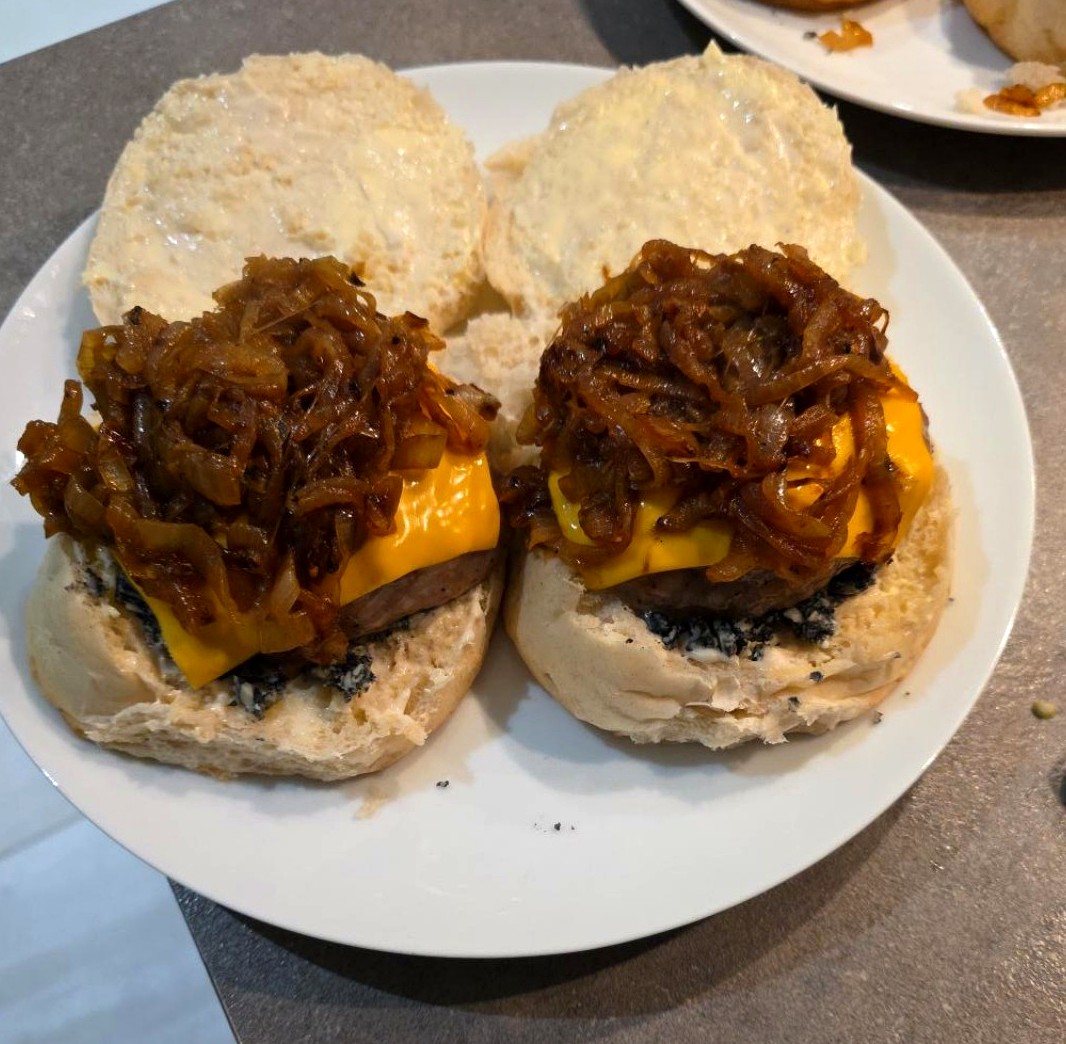 Two homemade pork burgers on Lancashire oven-bottom muffins, topped with melted cheese and dark caramelised onions on a white plate