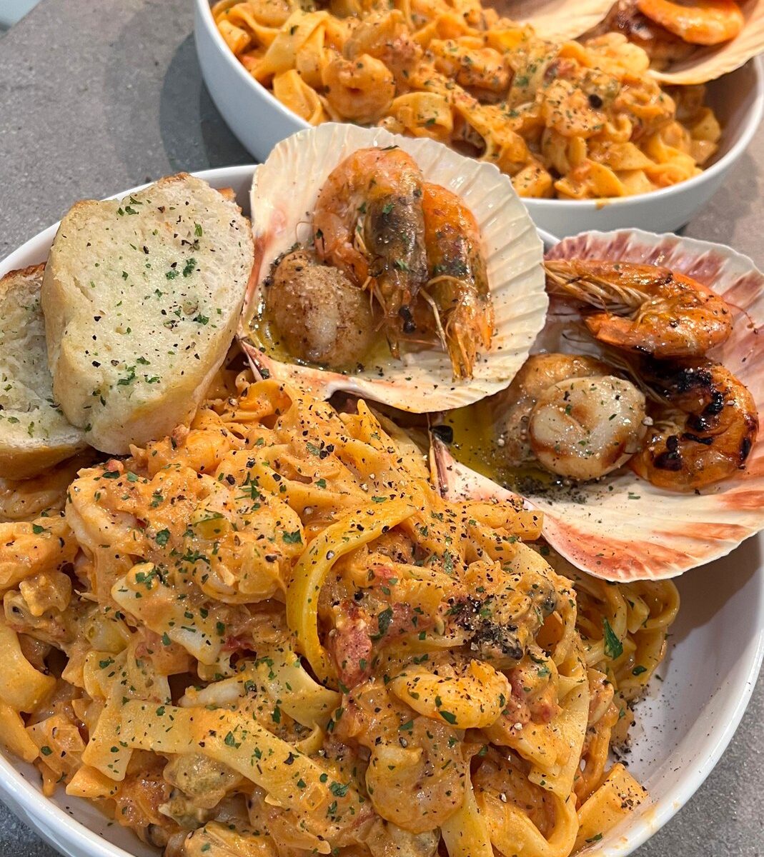 Two homemade bowls of creamy seafood tagliatelle with garlic bread, king scallops and shell-on prawns served in scallop shells